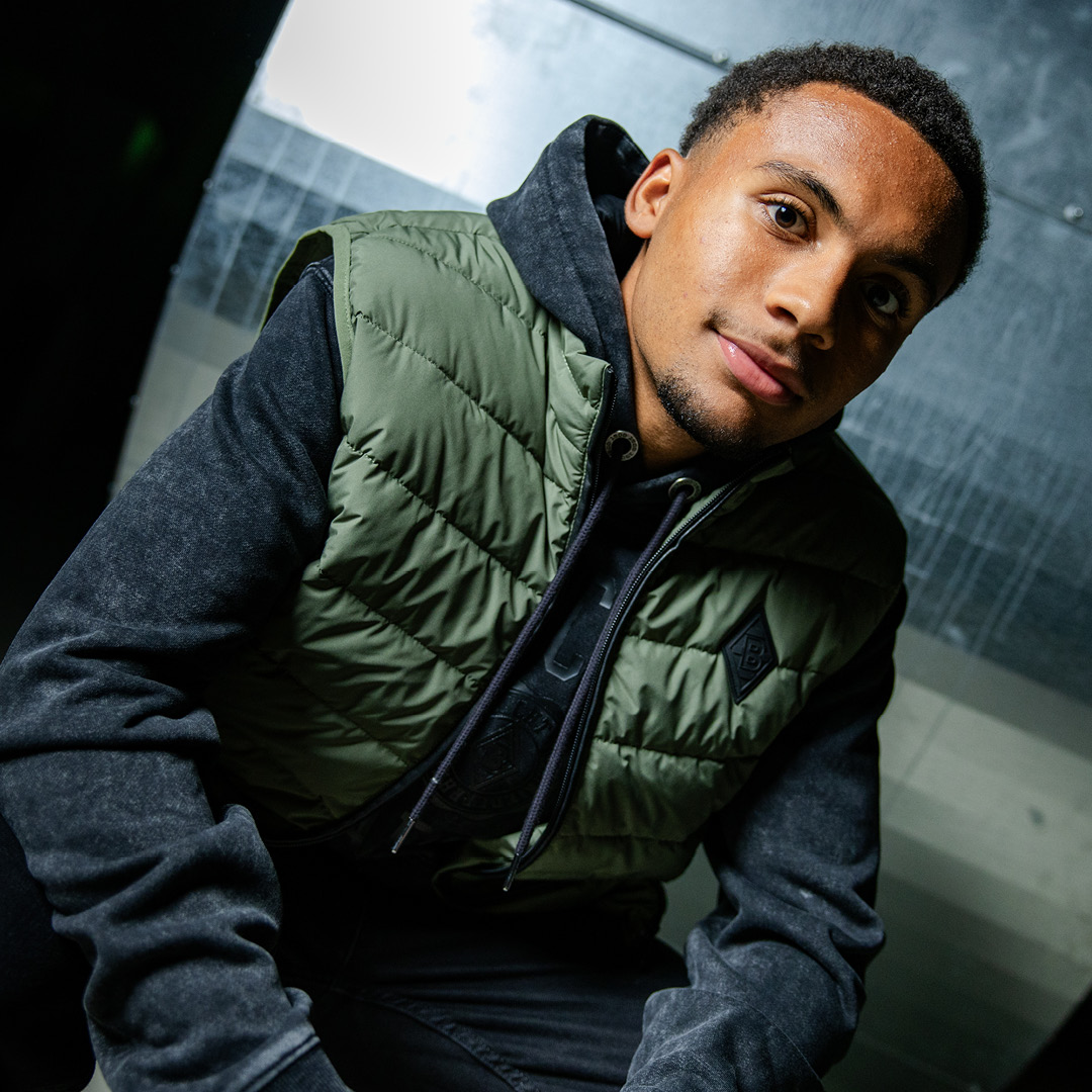 Man wearing a green quilted vest and black hoodie, sitting and looking towards the camera with a slight smile.