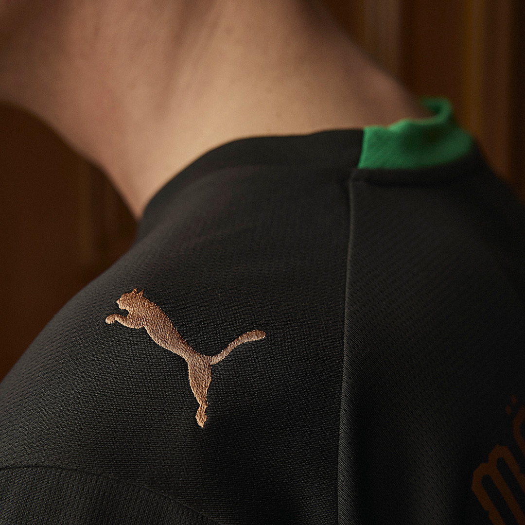 Close-up of a person wearing a black shirt with a gold embroidered Puma logo on the shoulder and a green collar detail.