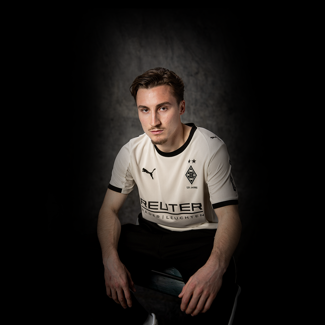 Eine Person im weißen Trikot von Borussia Mönchengladbach sitzt vor einem dunklen Hintergrund und schaut mit neutralem Blick in die Kamera.