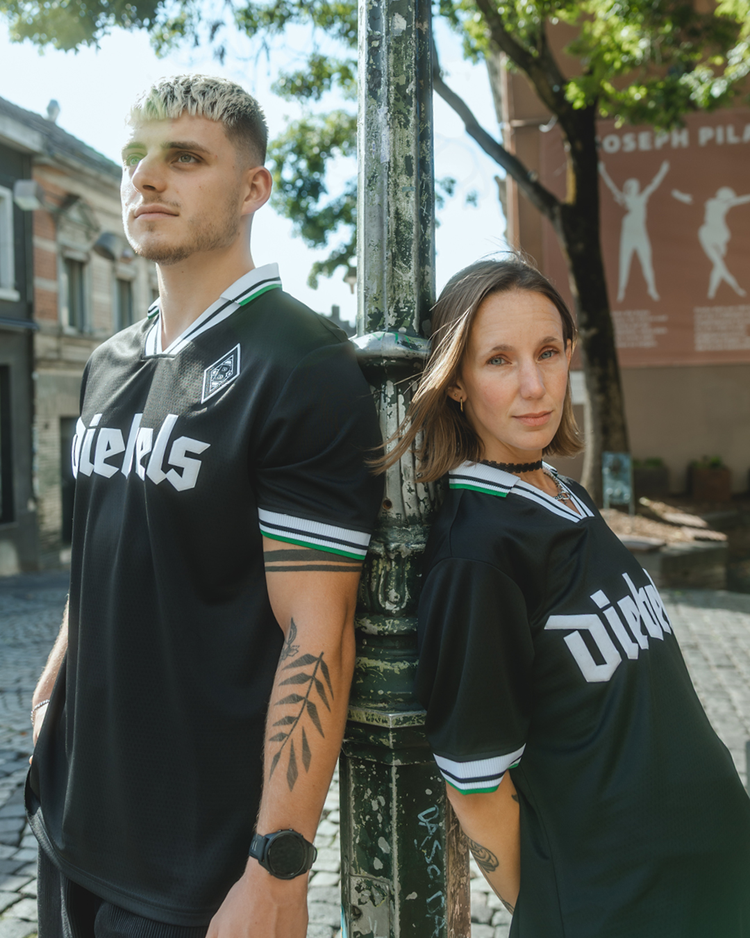 Two people in matching black jerseys with "DIESELS" text, leaning against a pole on a sunny street, with trees and buildings in the background.