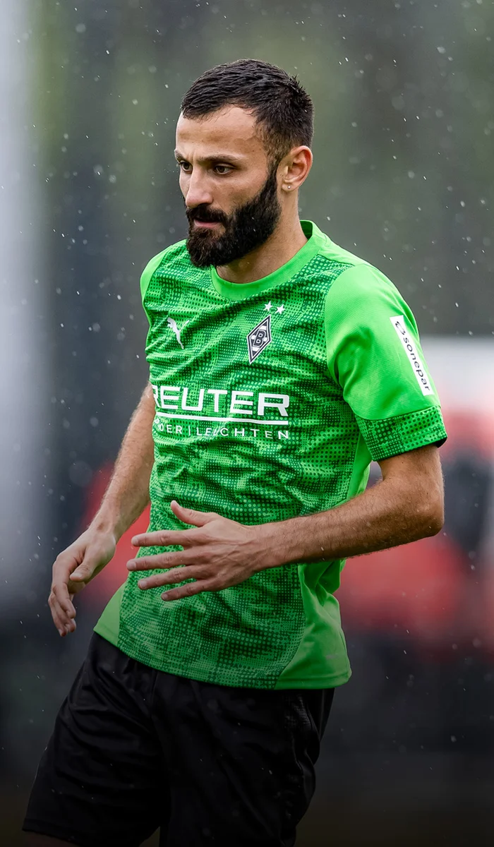 Soccer player in a green jersey with sponsor logos, playing in the rain on a blurred field background.