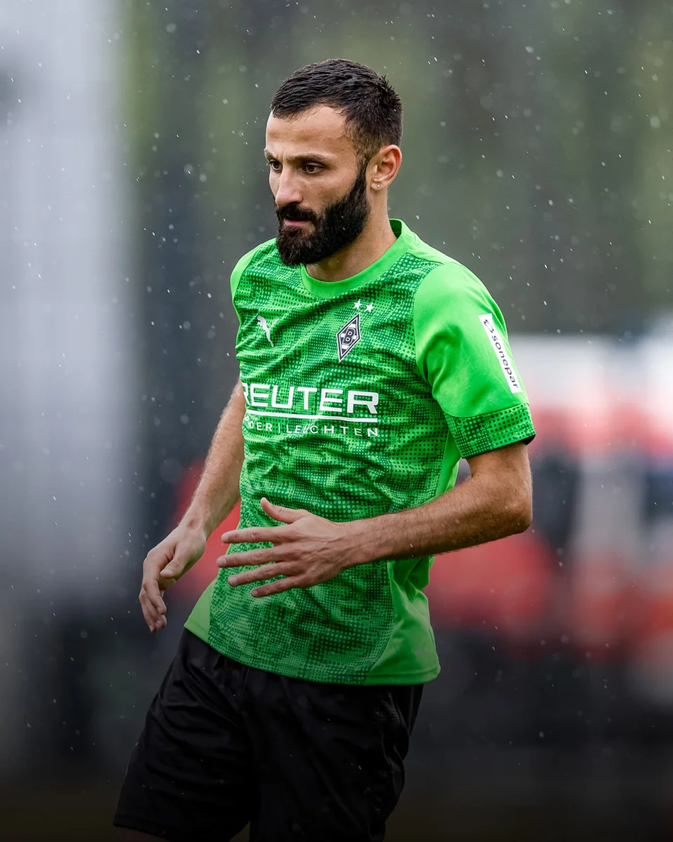Soccer player in a green jersey with sponsor logos, playing in the rain on a blurred field background.