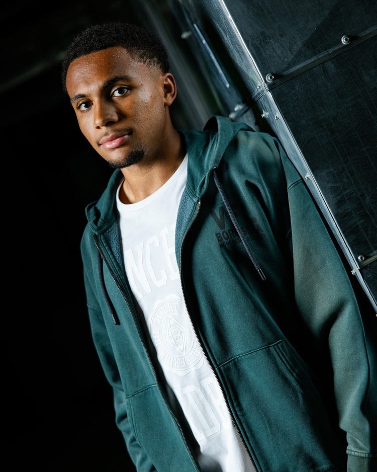 A man wearing a green hoodie and a white shirt stands against a metallic background, looking at the camera with a neutral expression.