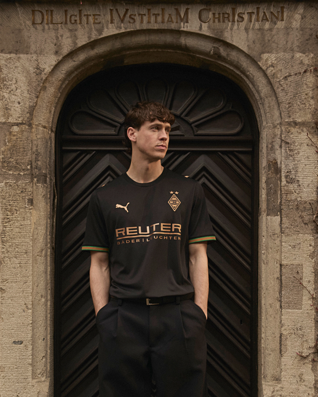 Man in a black football jersey stands in front of an ornate wooden door with Latin inscription above, arms in pockets.