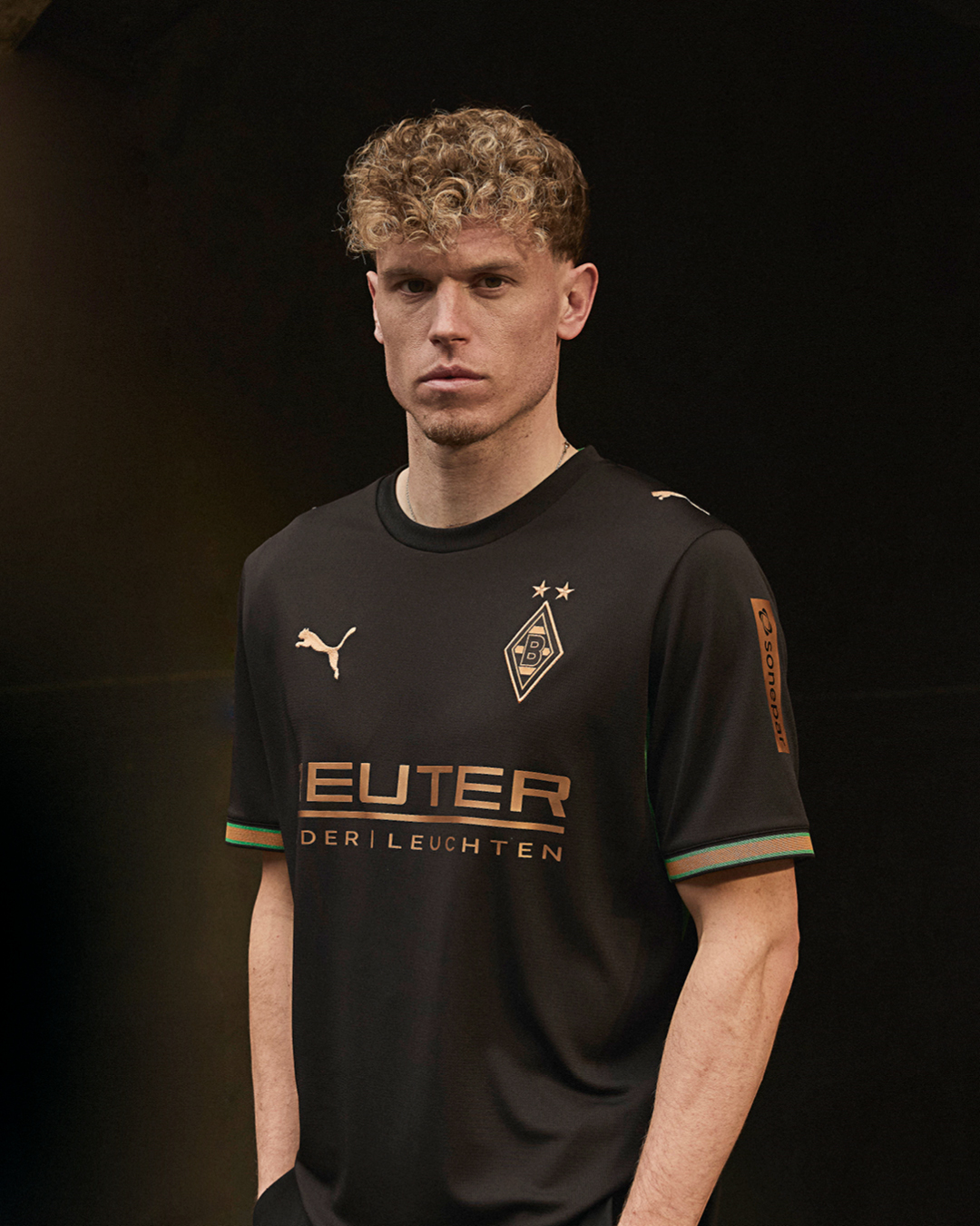 Man wearing a black Borussia Mönchengladbach football jersey with sponsor logos, standing against a dark background.