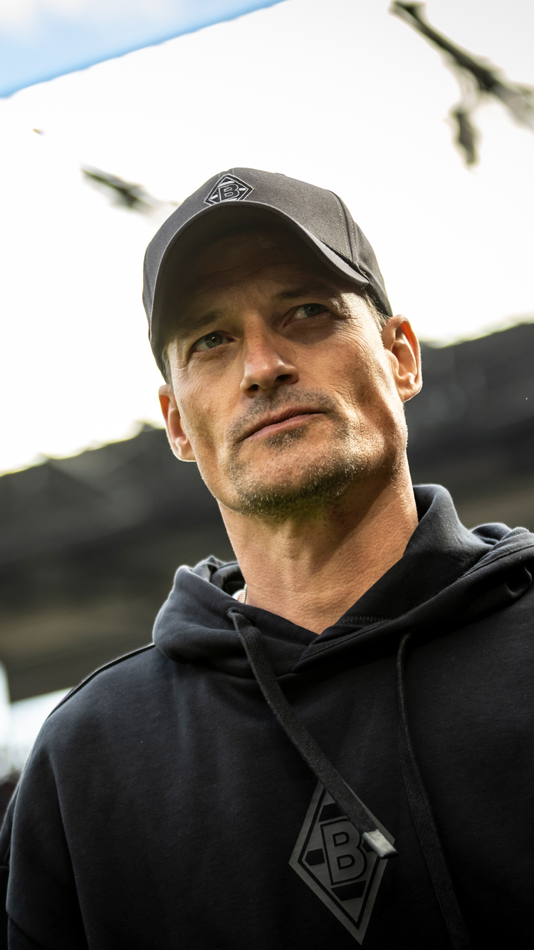 A person wearing a black cap and hoodie with a logo, standing outdoors with a thoughtful expression.