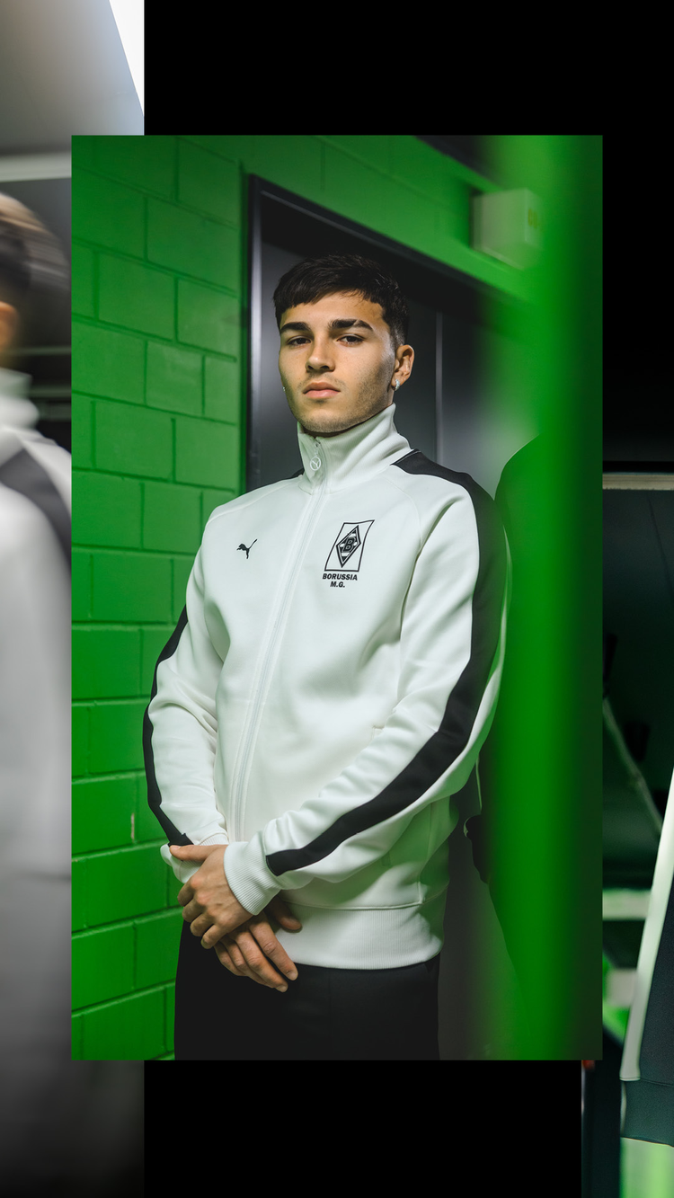 A person in a white and black Puma track jacket stands against a green tiled wall, with hands clasped in front.