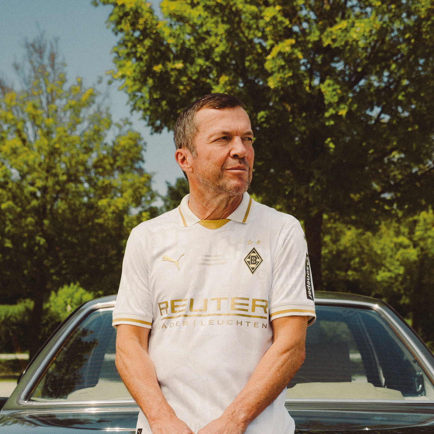 Man in a white sports jersey stands in front of a car, with trees in the background on a sunny day.