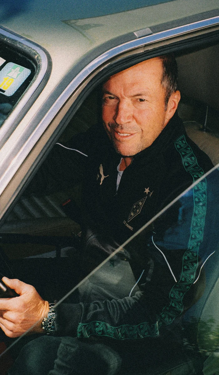 Ein Mann in einer Sportjacke sitzt in einem Auto und lächelt durch das offene Fenster in die Kamera