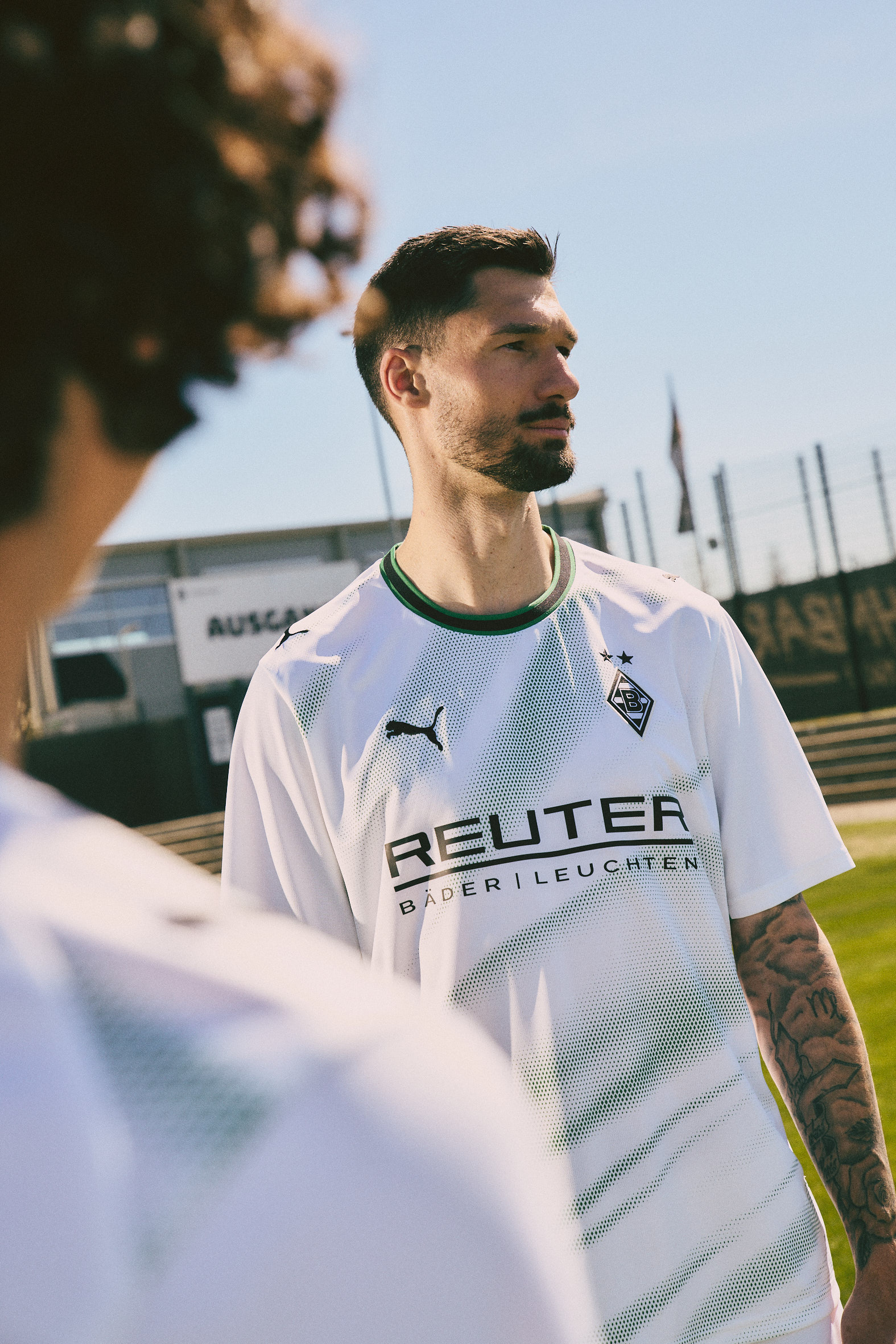 Soccer player in a white and green Puma jersey with "Reuter" logo, standing outdoors on a sunny day.