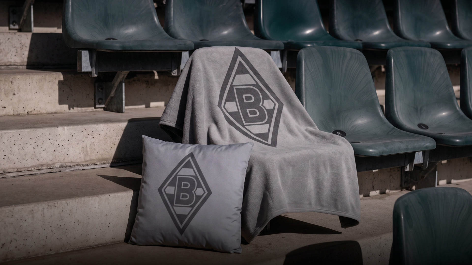 A gray blanket and pillow with a black geometric logo are placed on seating steps in an empty stadium.