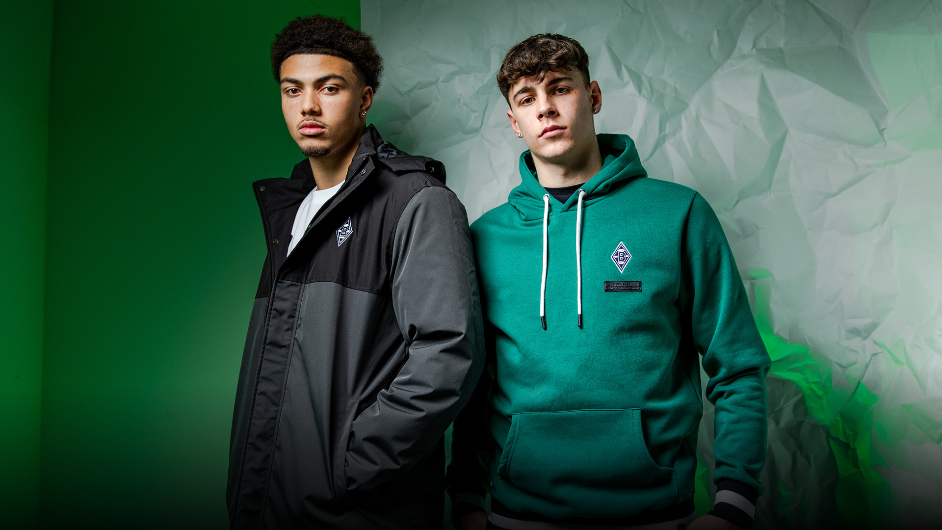 Two men in casual sportswear stand against a green and crumpled white background, exuding a relaxed and confident demeanor.