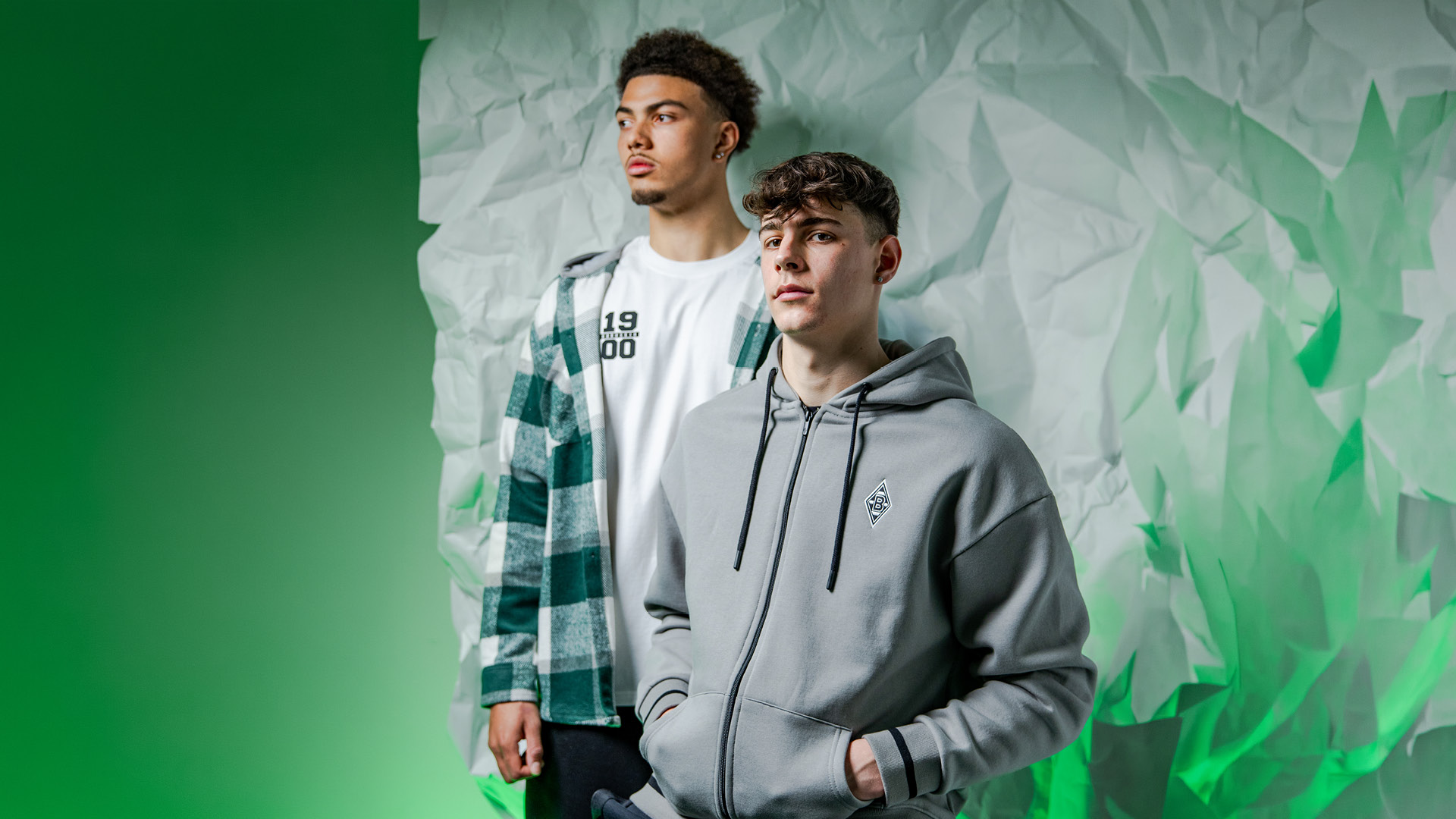 Two young men in stylish casual wear stand against a textured green and white backdrop, one in a plaid shirt and the other in a grey hoodie.