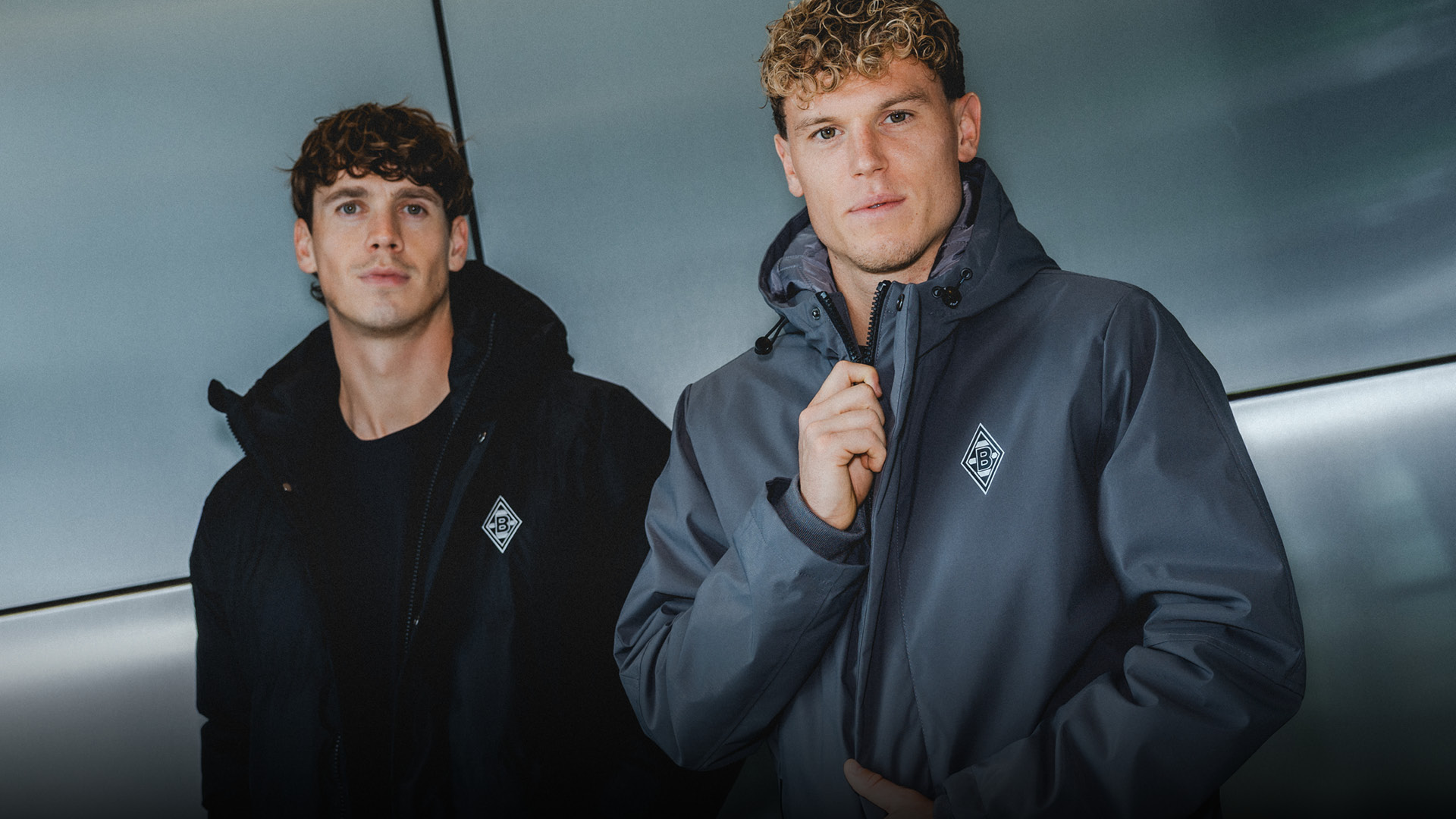 Two men in dark jackets stand indoors against a metallic backdrop, one zipping up his jacket while the other looks at the camera.