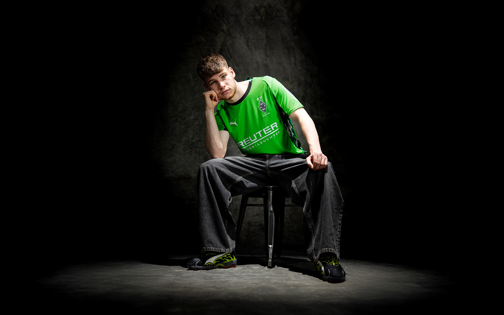 Person im grünen Trikot und weiten Jeans sitzt auf einem Hocker mit Kinn auf der Hand