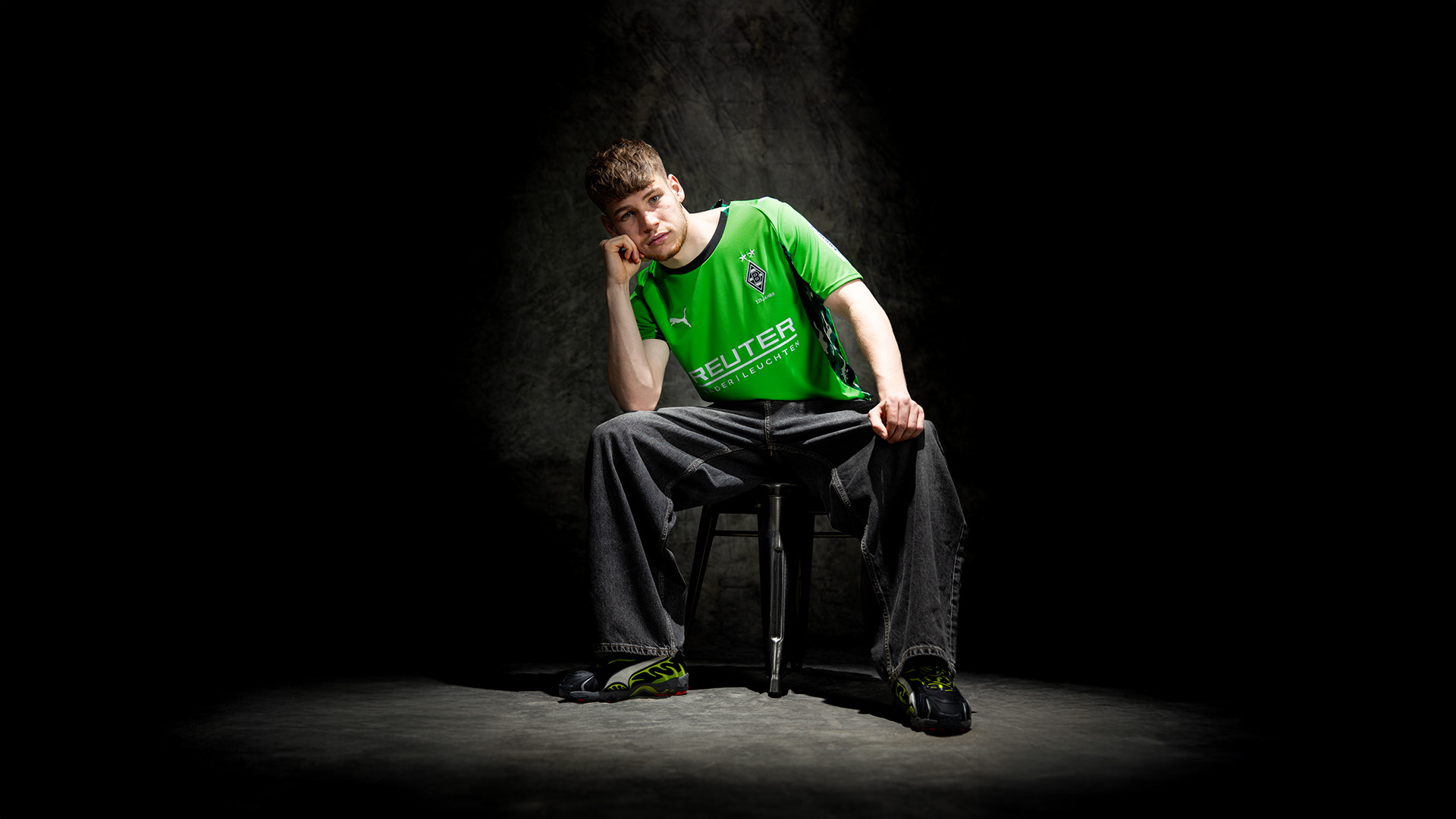 Person im grünen Trikot und weiten Jeans sitzt auf einem Hocker mit Kinn auf der Hand