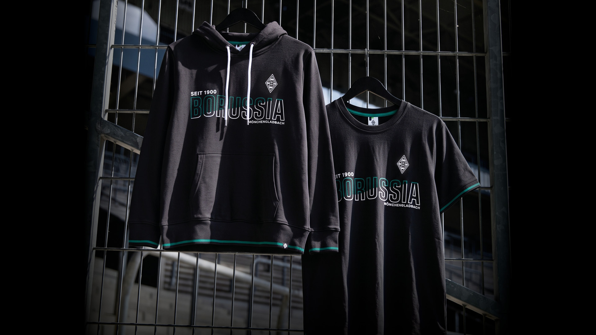 Black Borussia-themed hoodie and T-shirt with green accents hang on a metal fence, showcasing stylish sportswear.