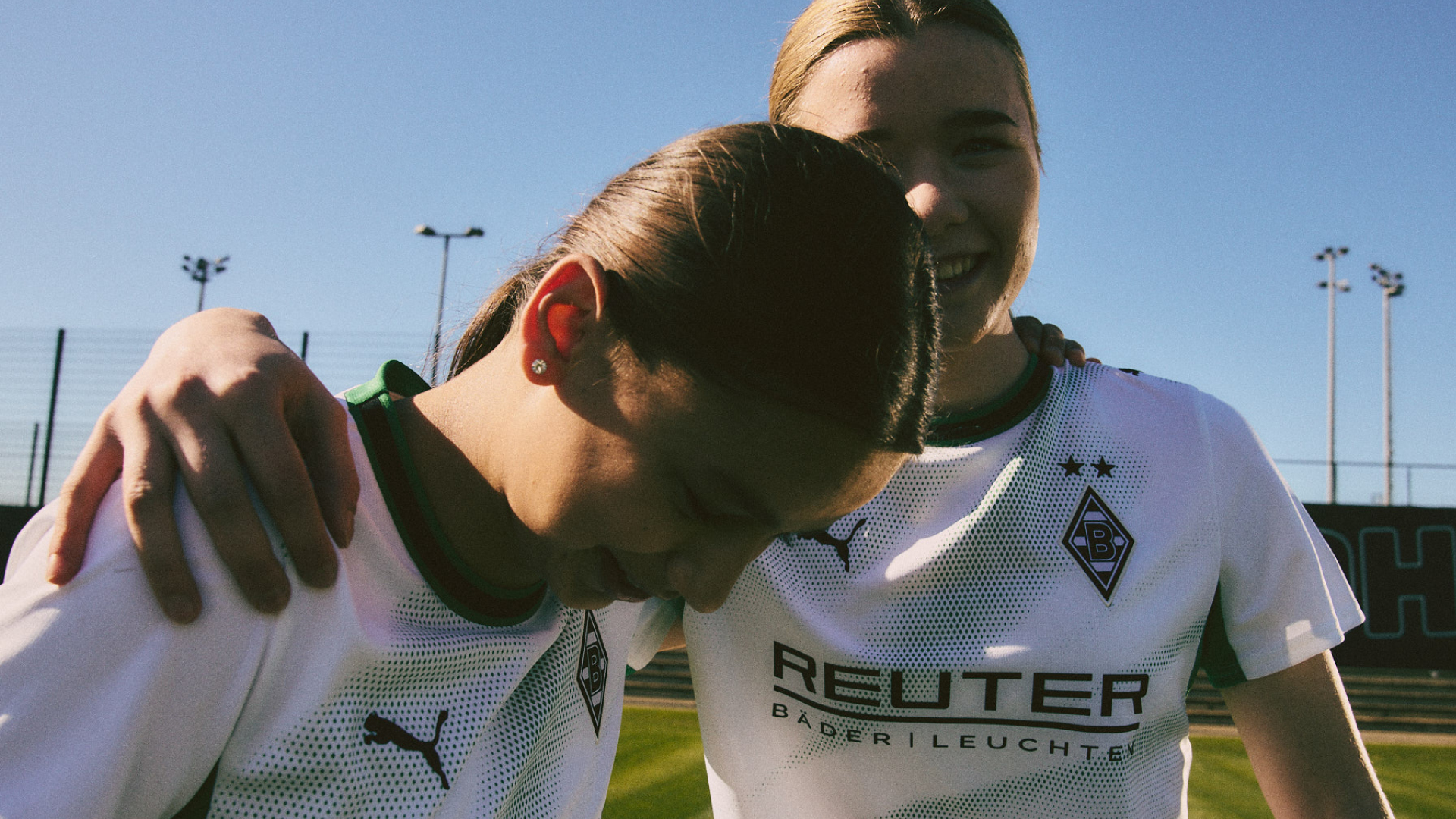 Two people in white sports jerseys embrace, smiling under a clear blue sky on a soccer field.