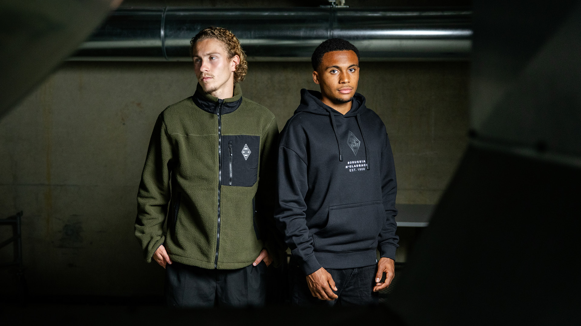 Two individuals standing in a dimly lit industrial setting, wearing casual hoodies and jackets, looking in different directions.