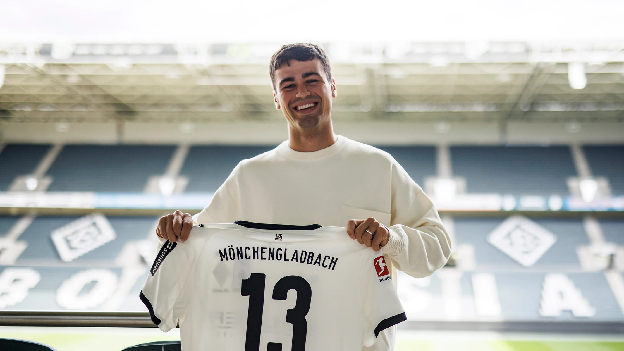 Lächelnde Person, die in einem Stadion ein Trikot von Borussia Mönchengladbach mit der Nummer 13 hält