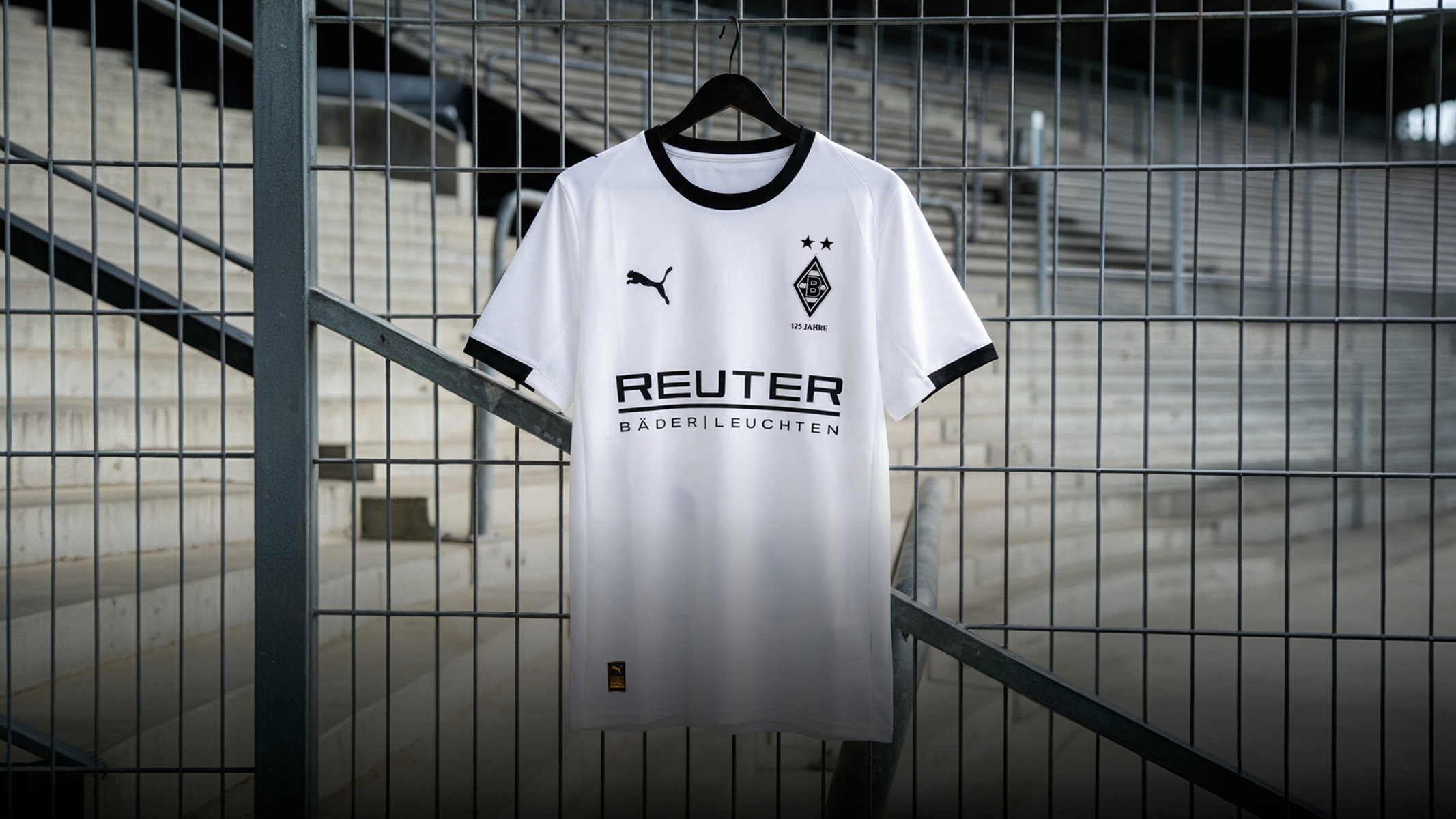 White soccer jersey with black trim, Puma logo, and "Reuter" text, hanging on a wire fence in an empty stadium.