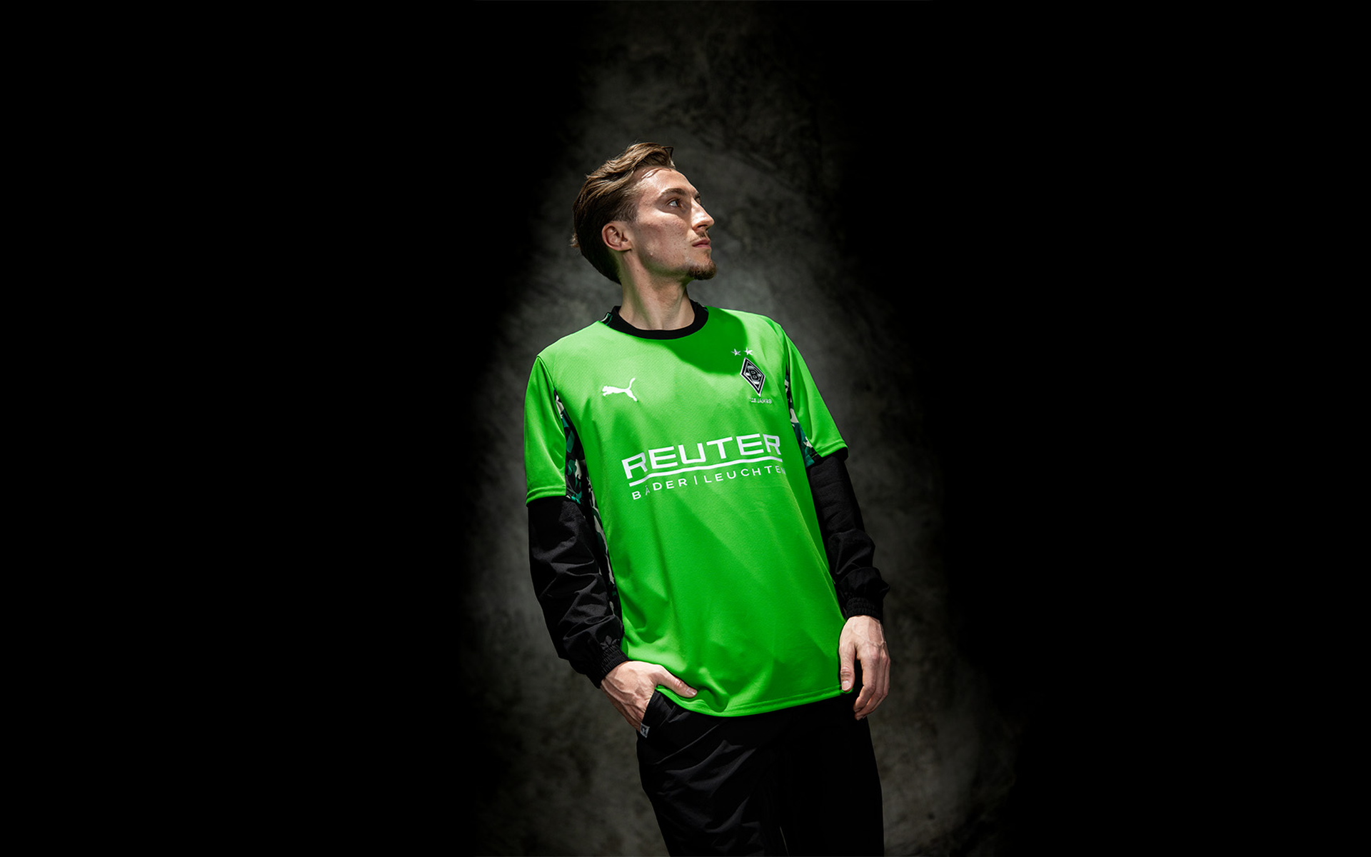 Eine Person in einem hellgrünen Sporttrikot steht vor einem dunklen Hintergrund und schaut mit den Händen in den Taschen zur Seite.