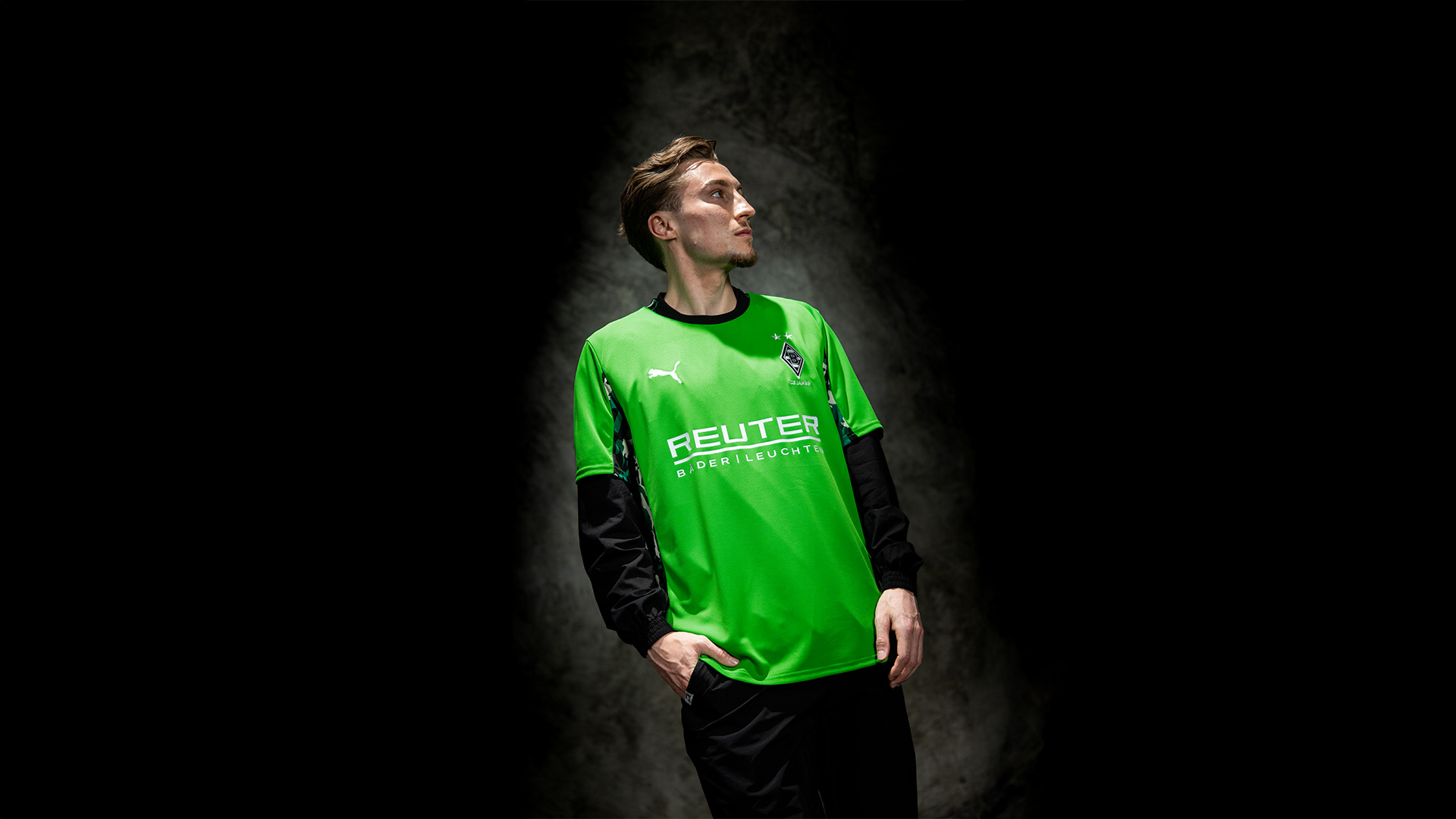 Eine Person in einem hellgrünen Sporttrikot steht vor einem dunklen Hintergrund und schaut mit den Händen in den Taschen zur Seite.