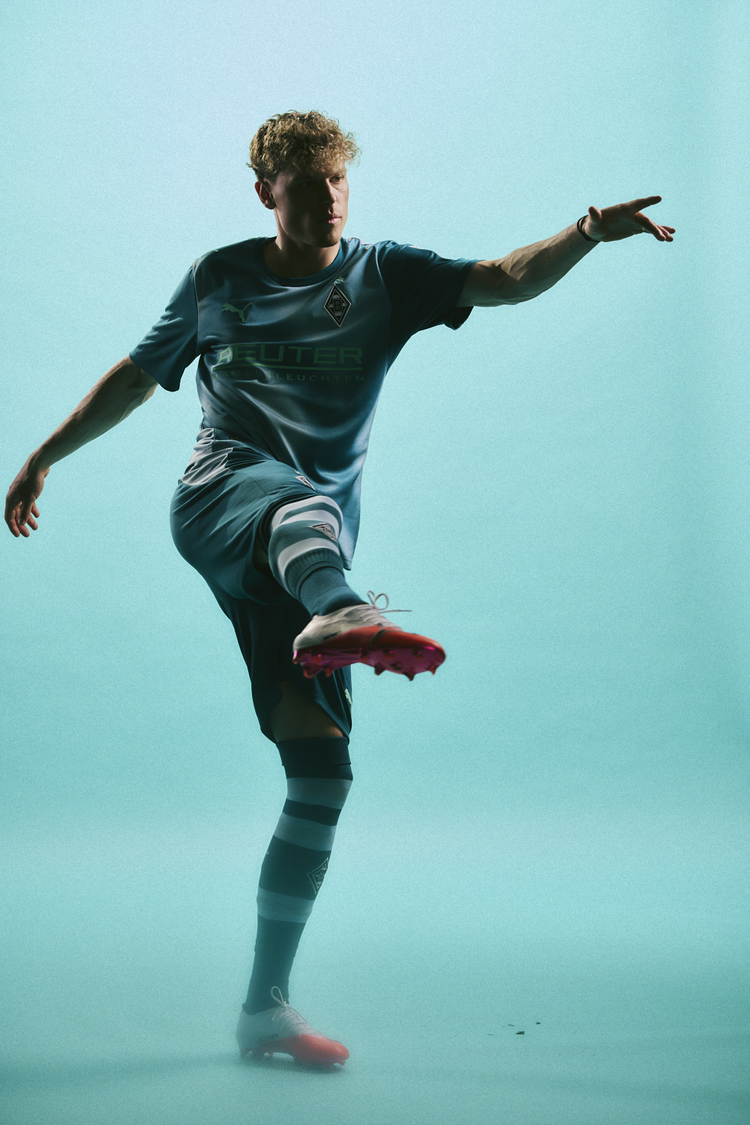 A man in a teal soccer uniform poses with one leg raised, wearing bright red cleats against a light teal background.