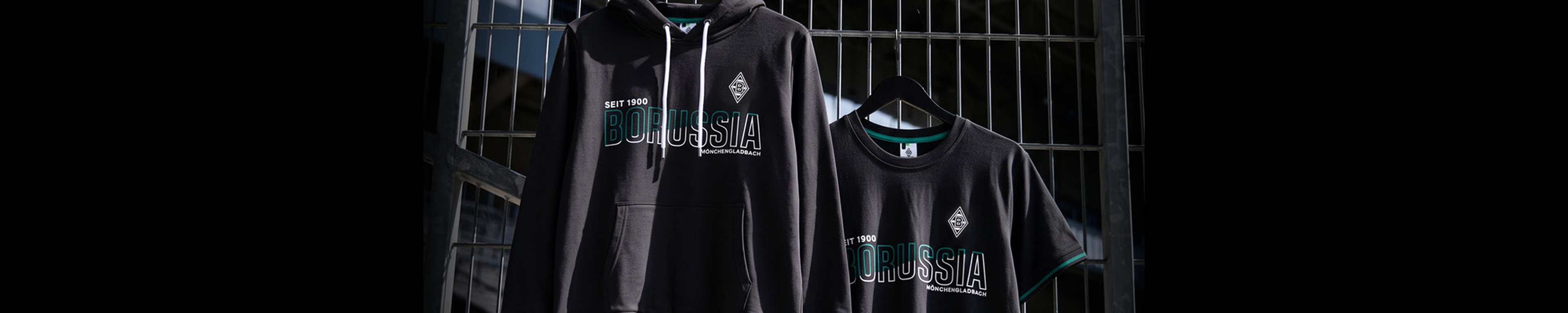 A black hoodie and T-shirt, both with logos, hang on a metal fence.
