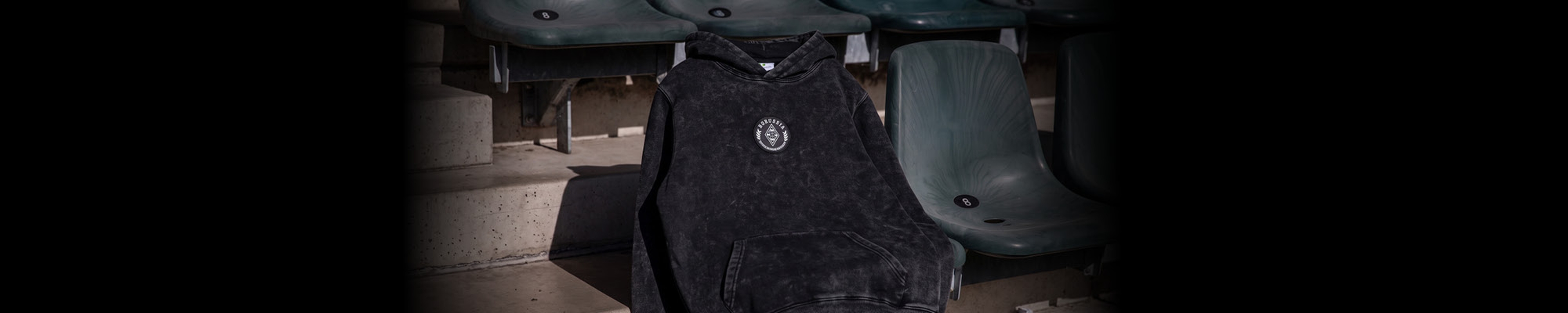A black hoodie with a small emblem draped over a green stadium seat under dim lighting.