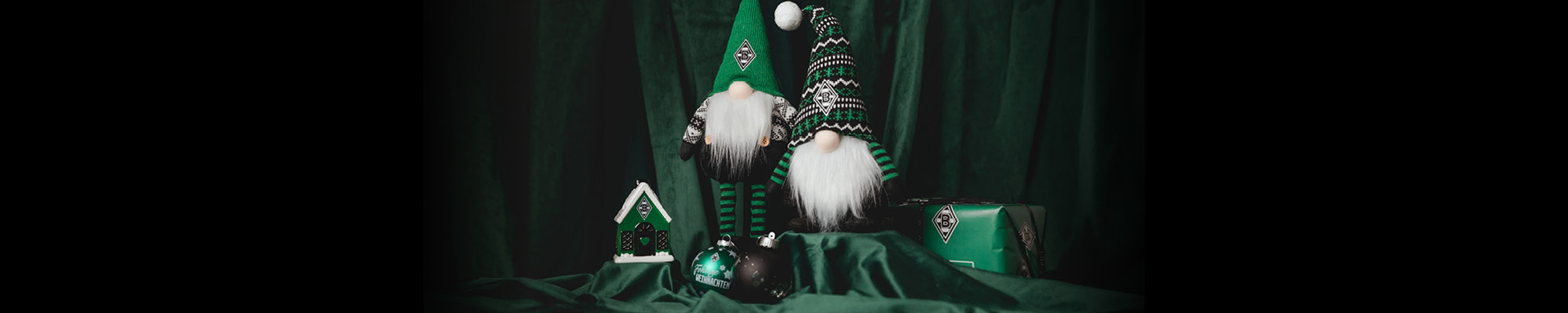 Two green gnomes with fuzzy beards sit among a birdhouse, ornaments, and a gift, all against a dark green draped background.