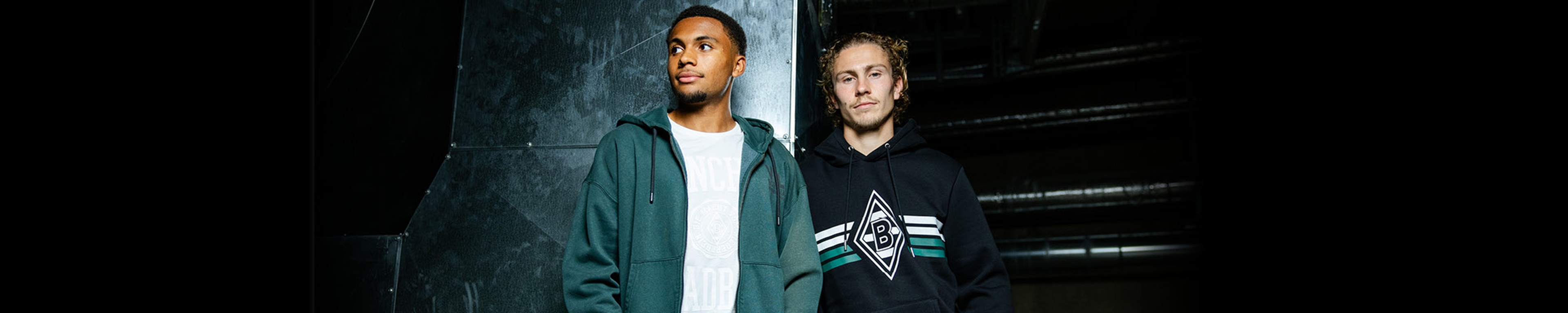 Two men standing side by side, one wearing a green jacket and the other a black hoodie, in a dimly lit industrial setting.