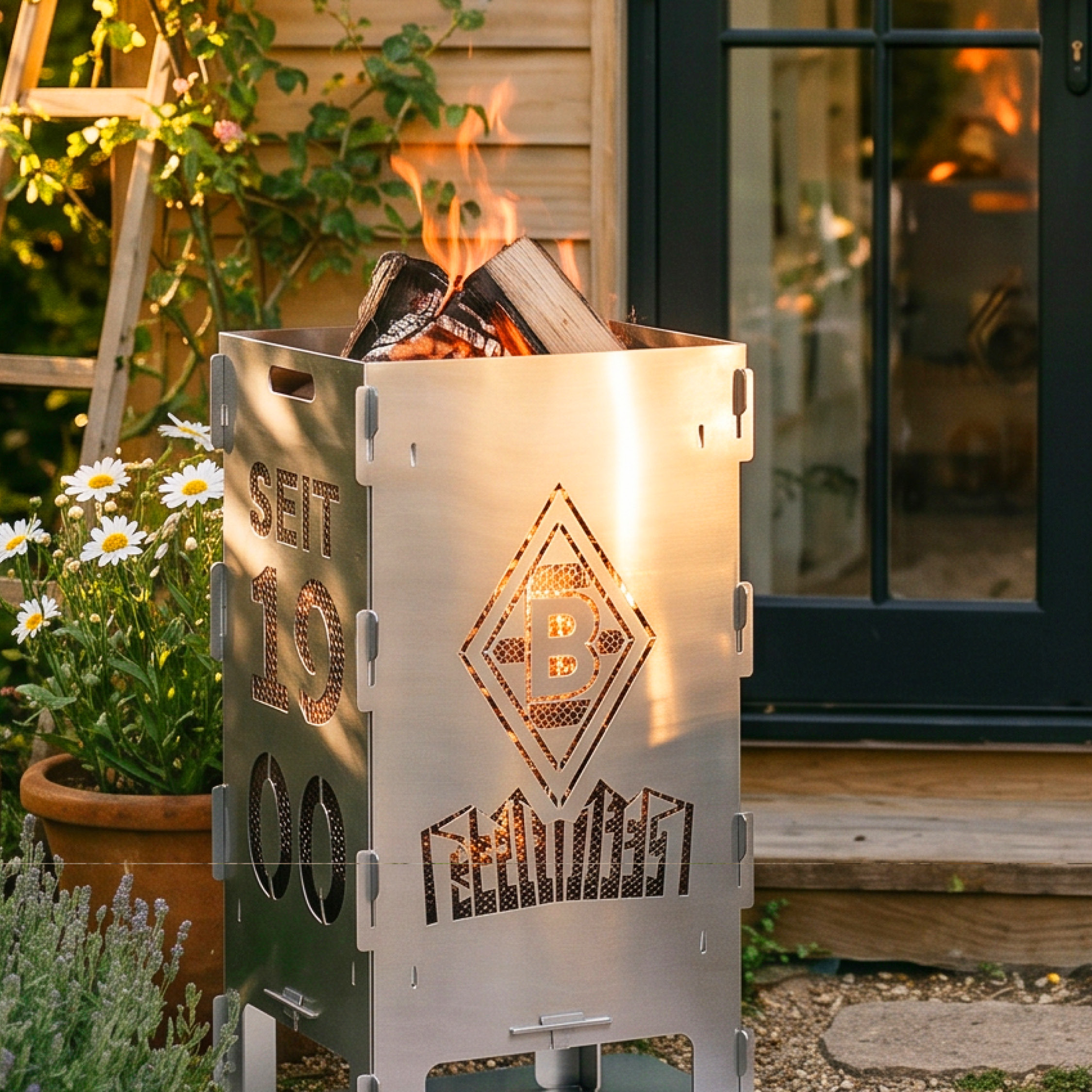 Metal fire pit with engraved design burning wood, in a garden with daisies. Black glass doors in the background reflect a warm glow.
