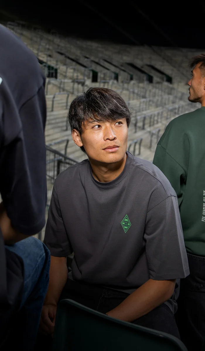 Three people in a stadium wearing dark green and gray shirts with unique designs, sitting and standing on steps.