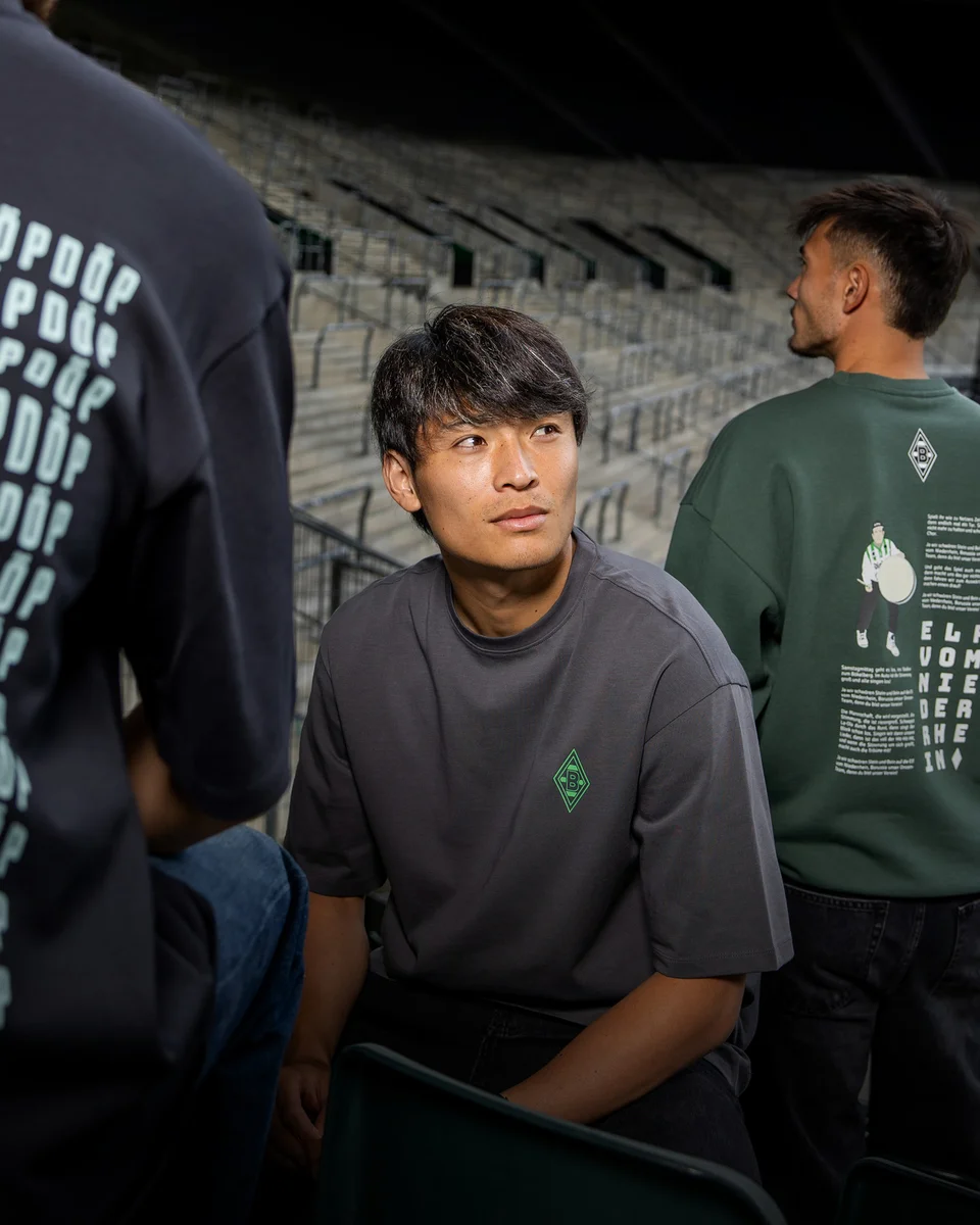Three people in a stadium wearing dark green and gray shirts with unique designs, sitting and standing on steps.