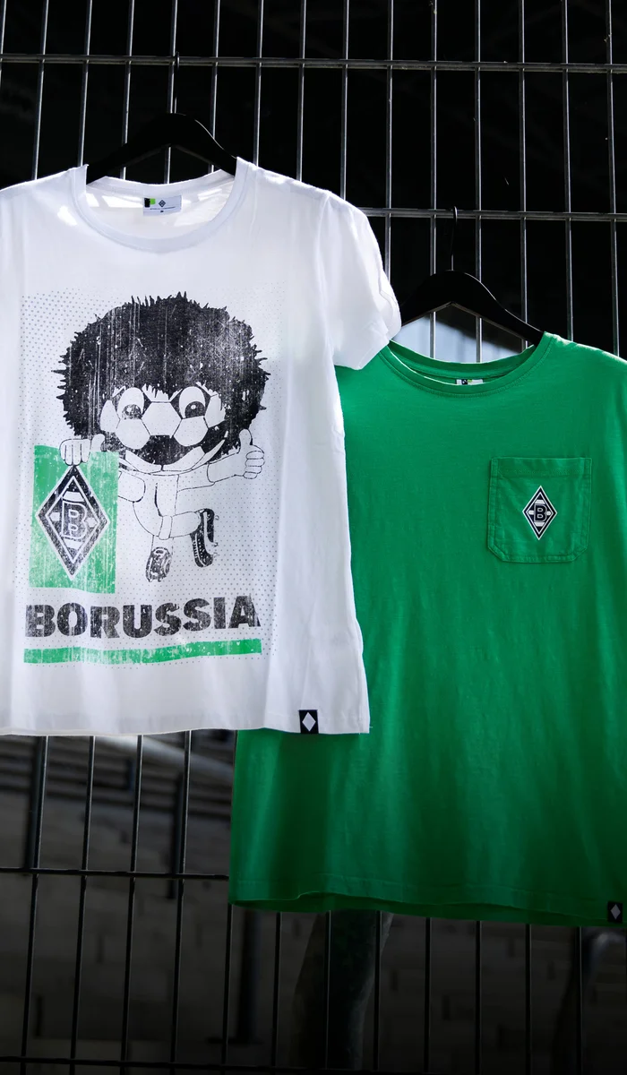 Two Borussia-themed T-shirts hang on a metal fence; one white with a cartoon design, the other green with a pocket and logo.
