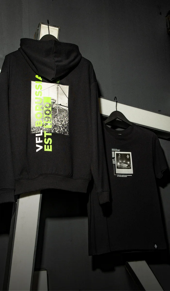 Black hoodie and T-shirt with graphic designs hang on a dark, industrial-style wall with white, angled beams.