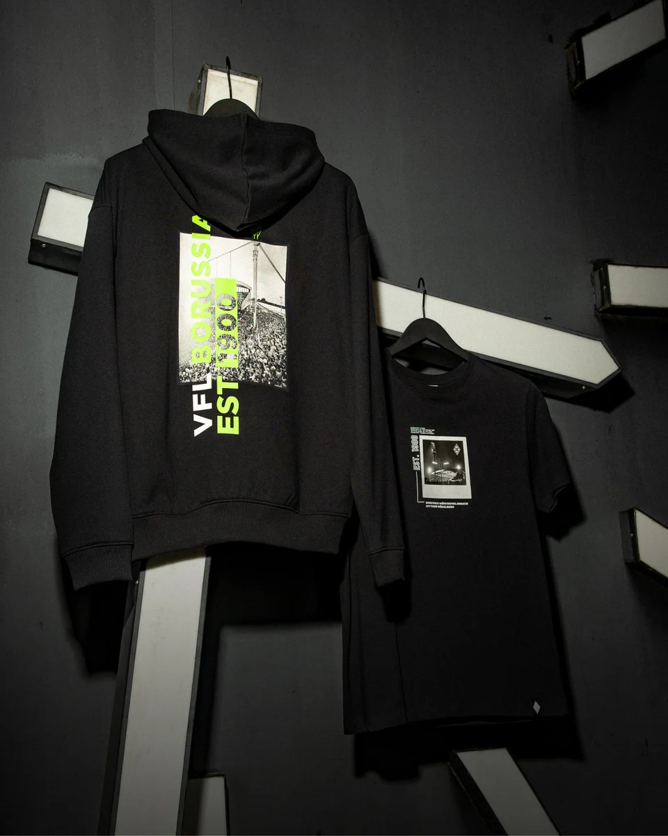 Black hoodie and T-shirt with graphic designs hang on a dark, industrial-style wall with white, angled beams.