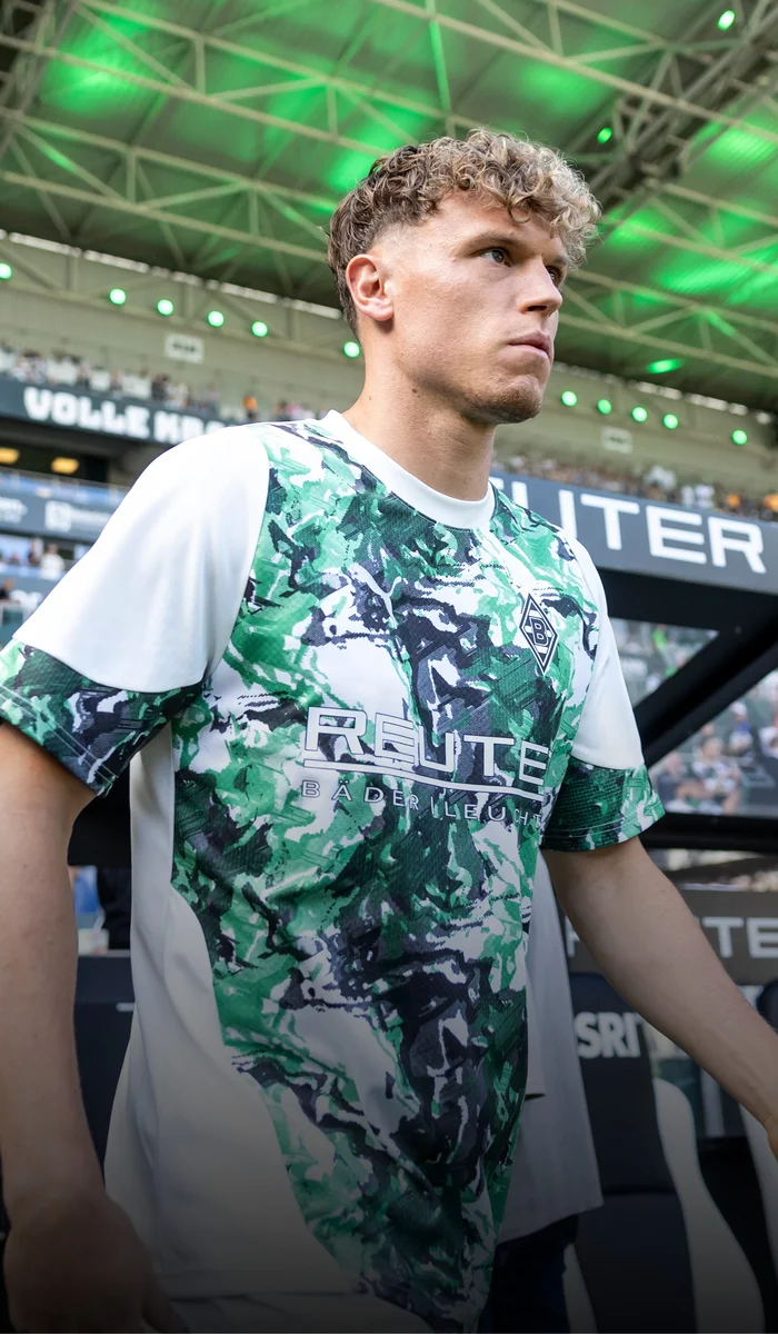 Soccer player in a green and white camo jersey walks onto the field under a stadium roof with green lighting.