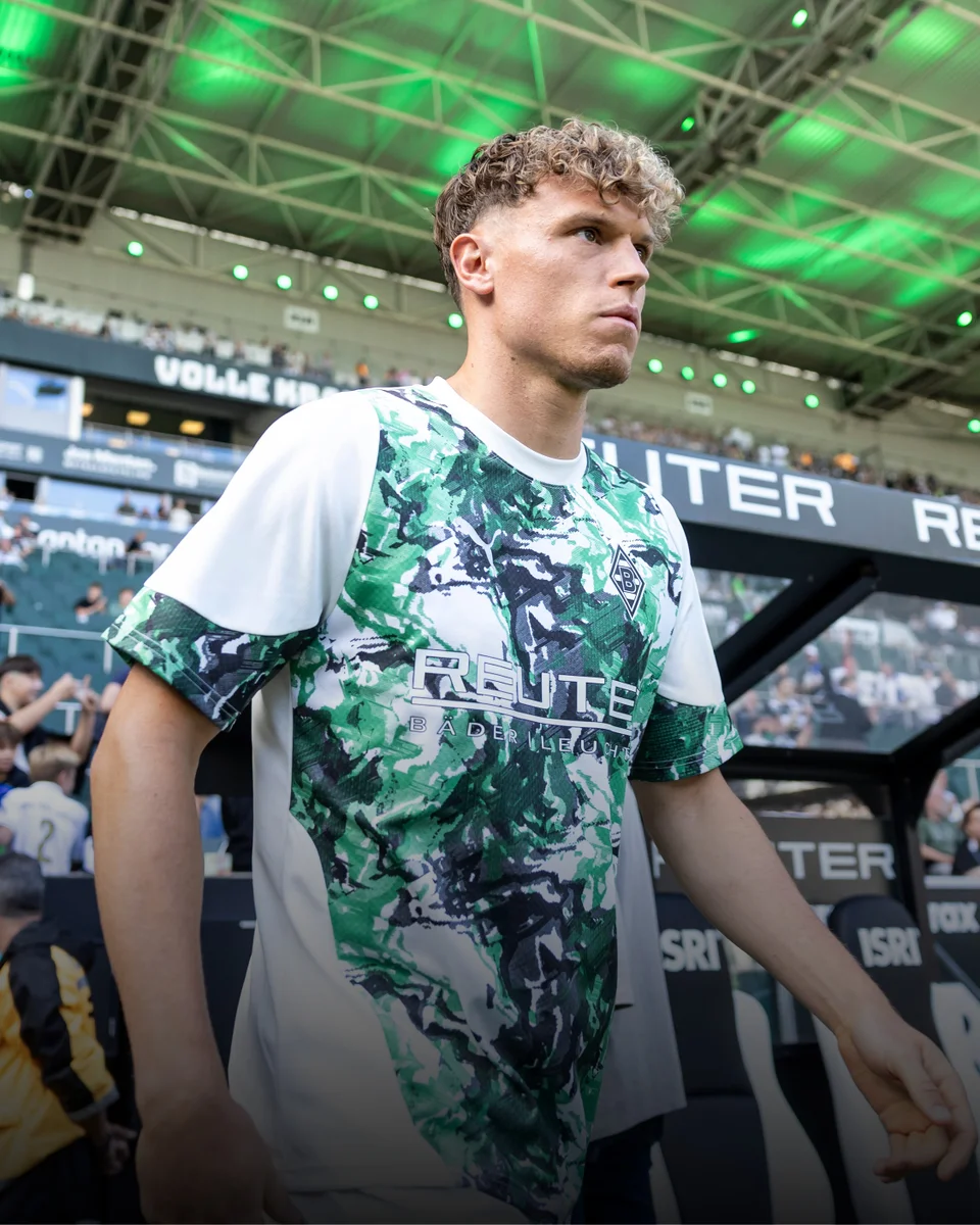 Soccer player in a green and white camo jersey walks onto the field under a stadium roof with green lighting.