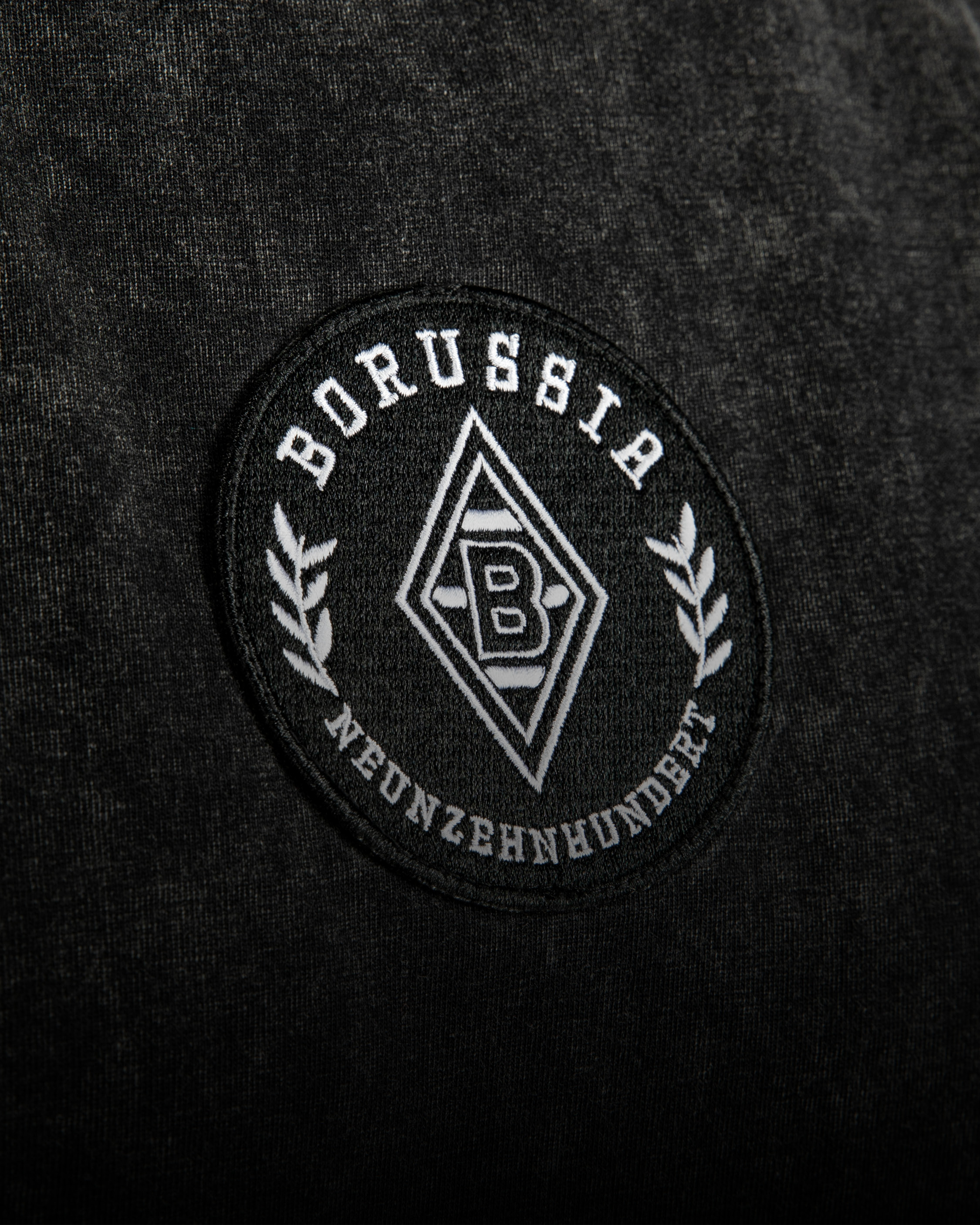 A Borussia Mönchengladbach emblem is embroidered on dark fabric, featuring a stylized "B" and laurel design with the word "Neunzehnhundert."