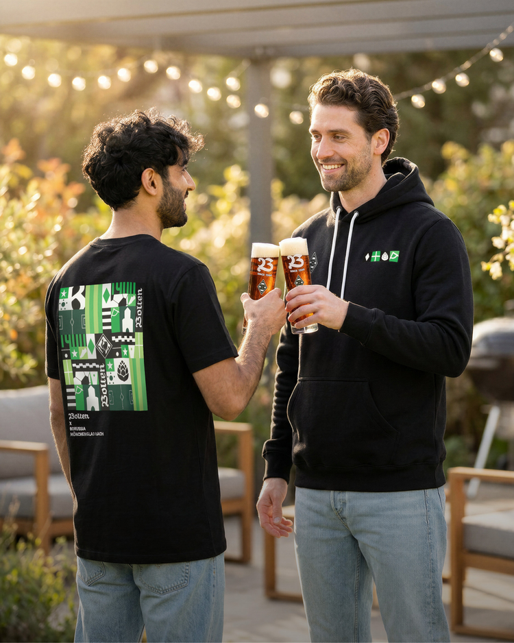 Two men smiling and toasting with beers in a sunny outdoor setting, wearing casual black outfits with a green-themed design.