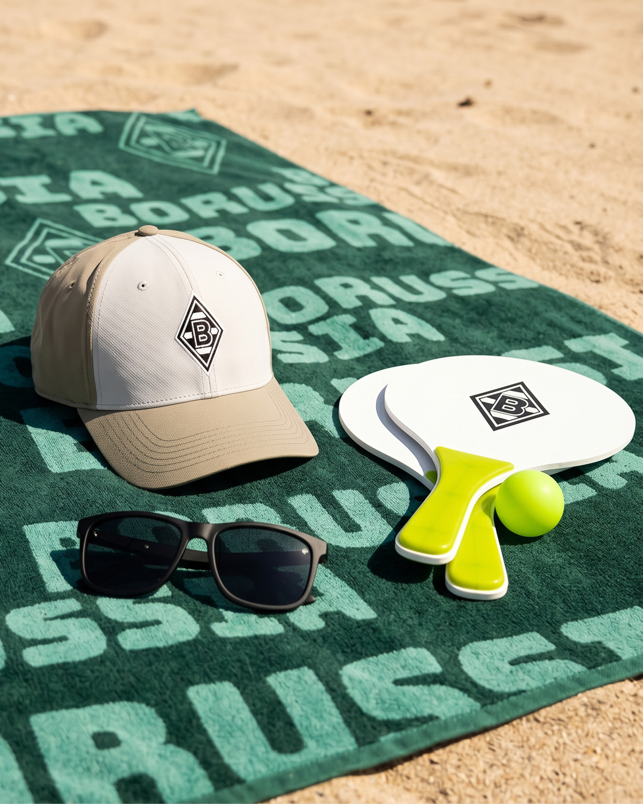 Beach setup with sunglasses, a cap, and ping pong paddles on a green "Borussia" towel on sand.