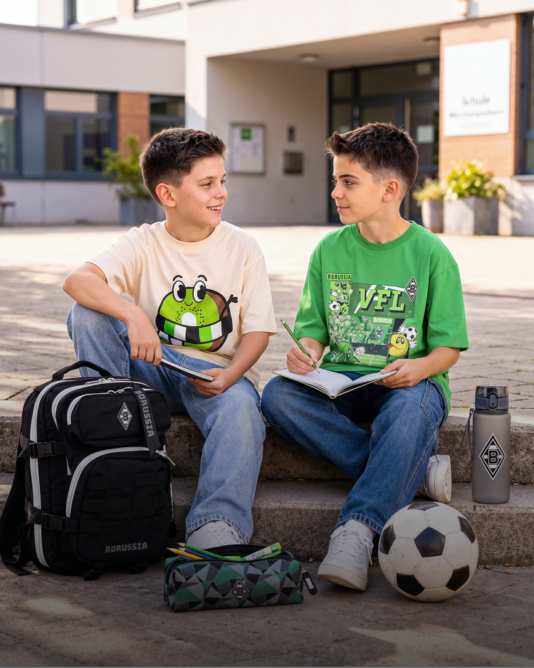 Two boys sitting on steps with backpacks and a soccer ball, wearing graphic t-shirts, talking while one writes in a notebook.