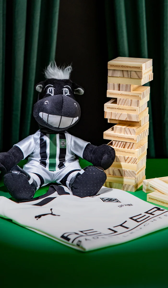 A stuffed horse toy in a soccer jersey sits next to a Jenga tower and scattered blocks, with a folded jersey in the foreground on a green surface.