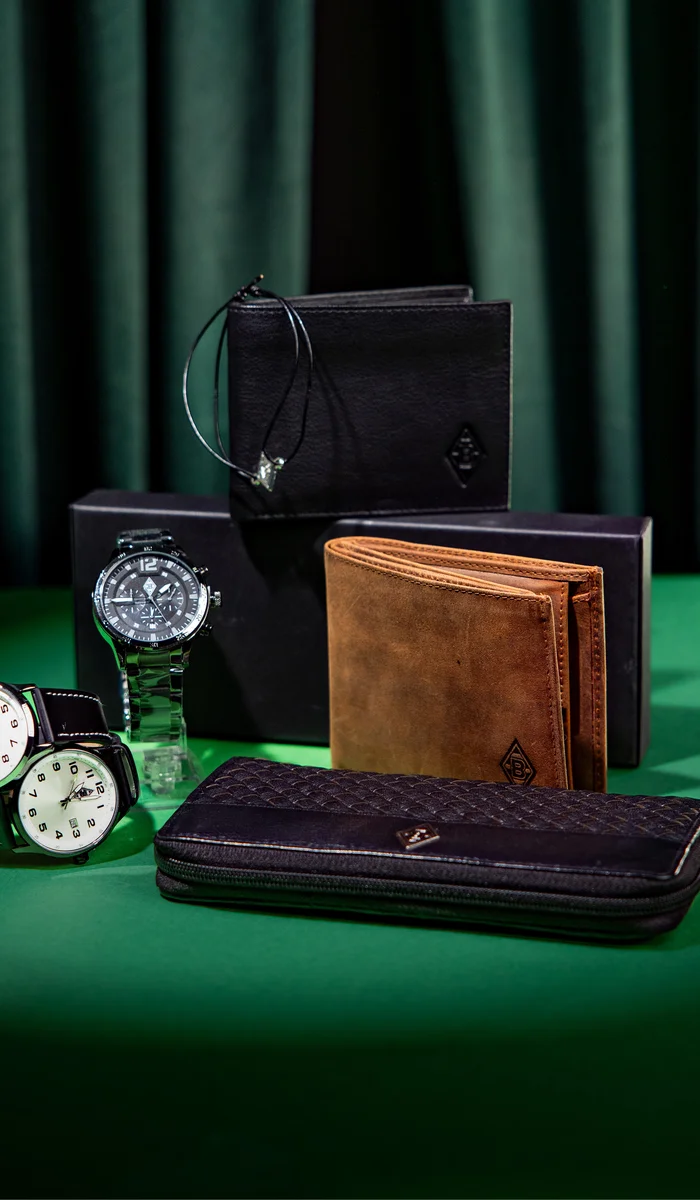Display of luxury accessories: two watches, a brown wallet, a black zippered wallet, and black boxes on a green surface with a curtain backdrop.