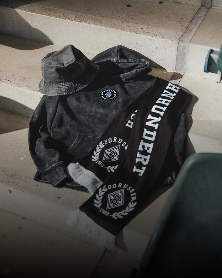 A black hoodie, bucket hat, and scarf with "Borussia" and "Jahrhundert" printed on them, lying on concrete steps in natural light.