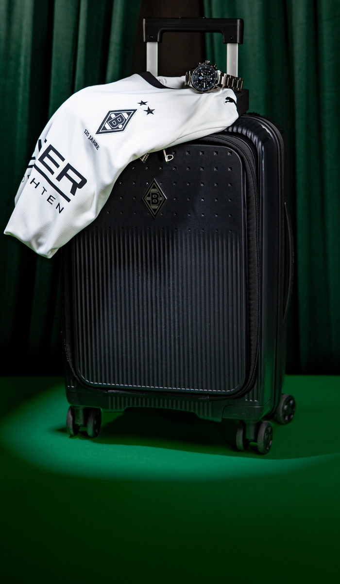 Black suitcase with a white sports jersey draped over it, featuring a logo and stars. A watch is placed on top. Green curtain background.