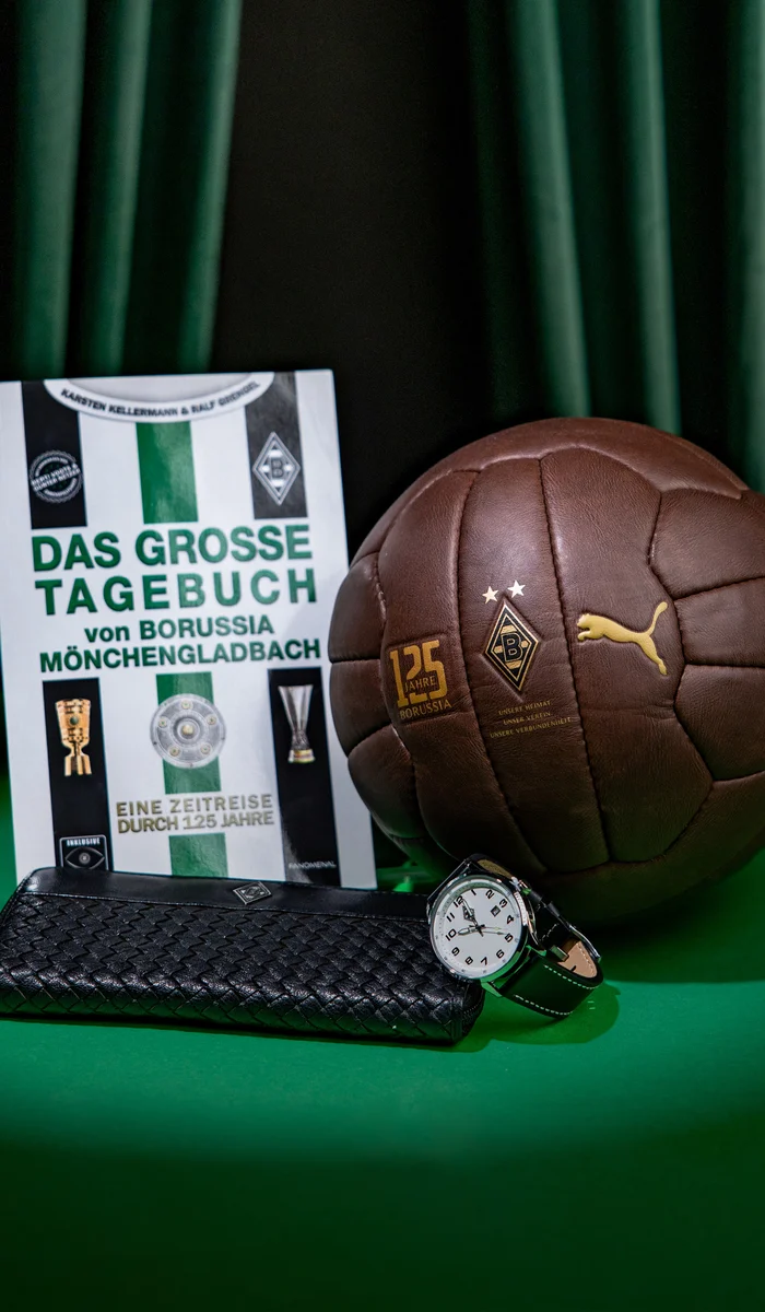 Book titled "Das Grosse Tagebuch von Borussia Mönchengladbach," a vintage-style soccer ball, a wristwatch, and a black pouch on a green surface.
