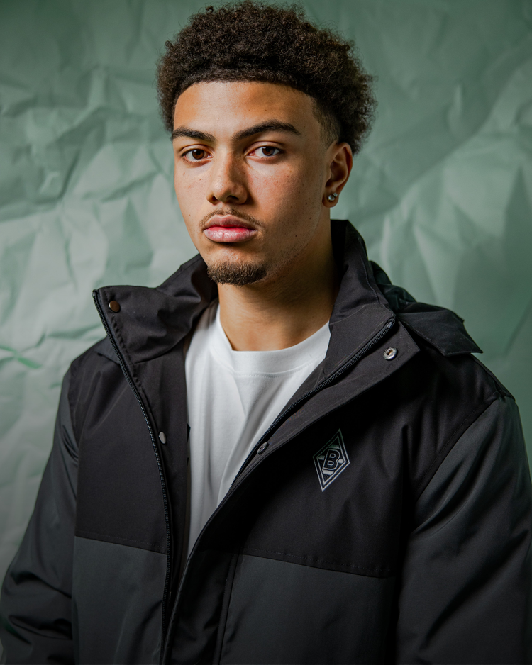 Young man with short curly hair wearing a black jacket over a white shirt, standing against a crumpled green backdrop, looking forward.